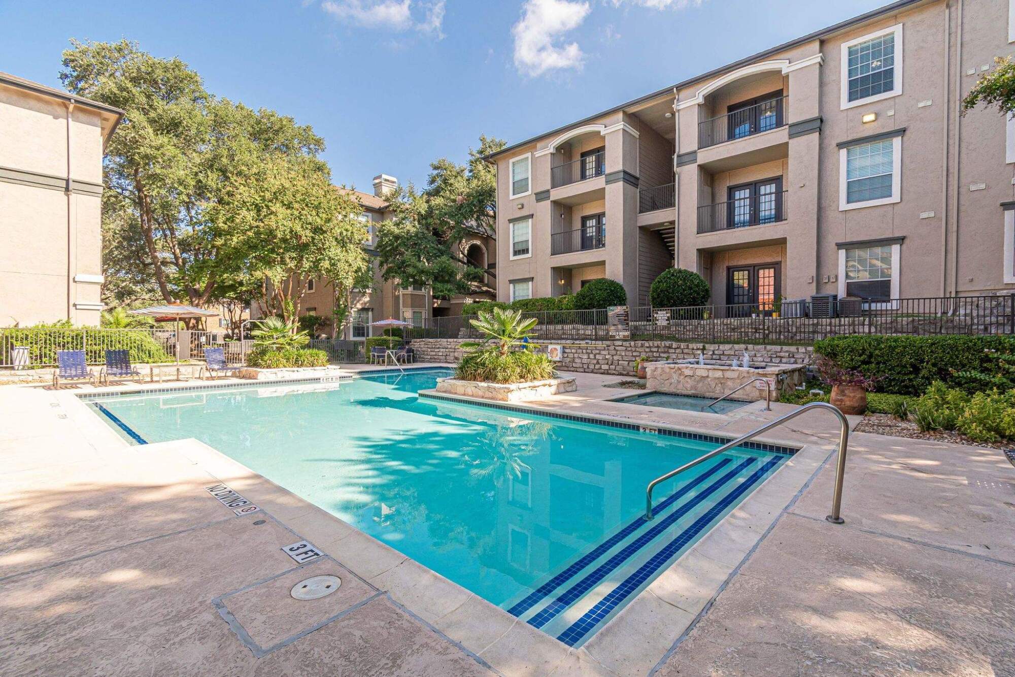 Outdoor swimming pool in front of a three-story apartment building with balconies and surrounding greenery.