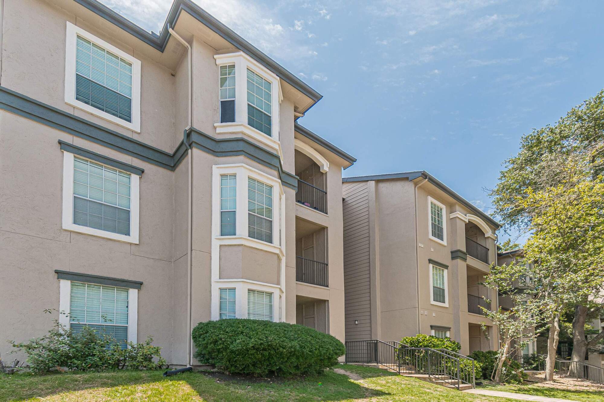 Three-story beige apartment building with white trim, large windows, and landscaped greenery under a blue sky.